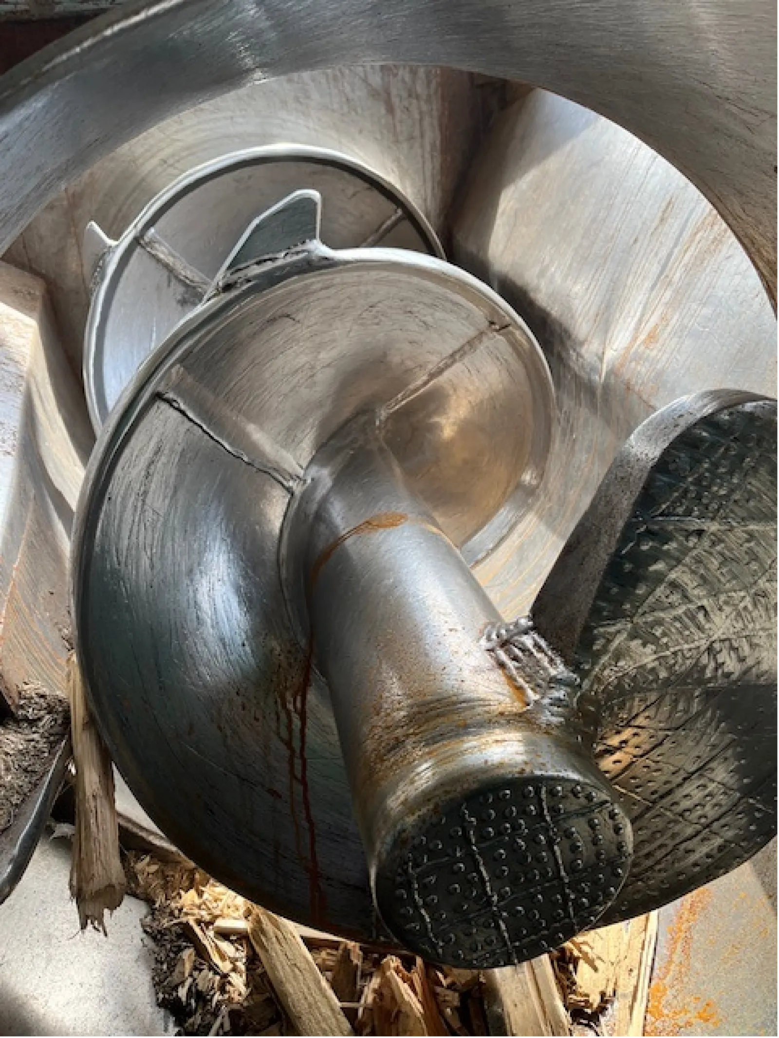 Close-up of a large metallic industrial screw conveyor with rust marks and wood debris nearby.