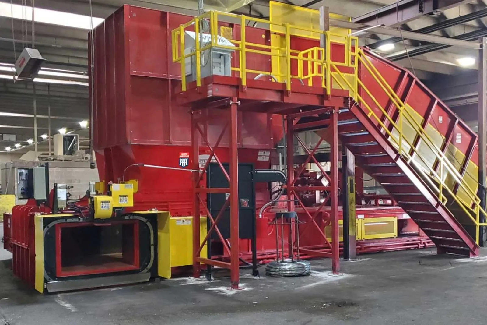 Large industrial metal shredder machine in a factory with red and yellow safety railings and stairs
