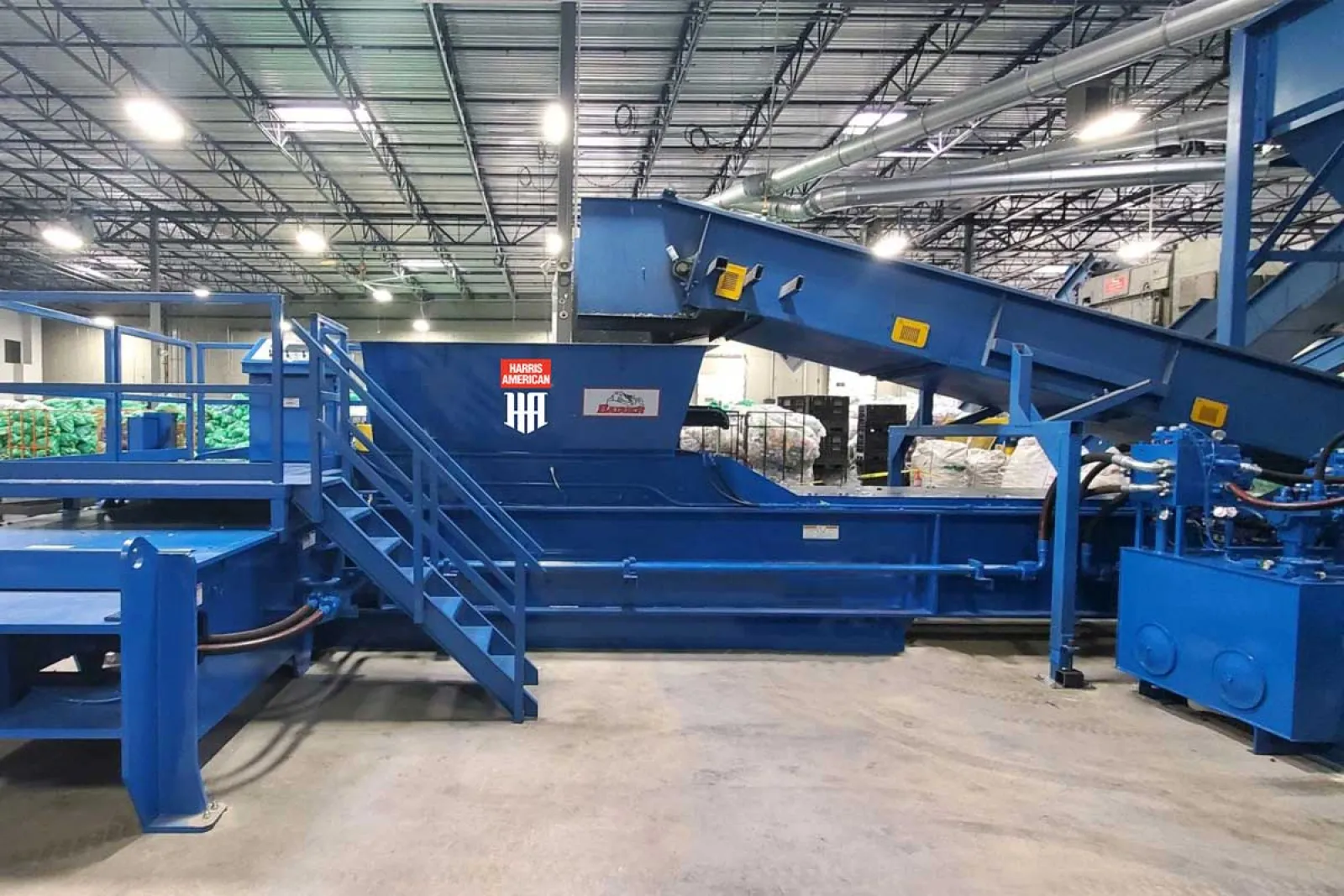 Large blue industrial conveyor system inside a warehouse with stairs and machinery under bright lighting