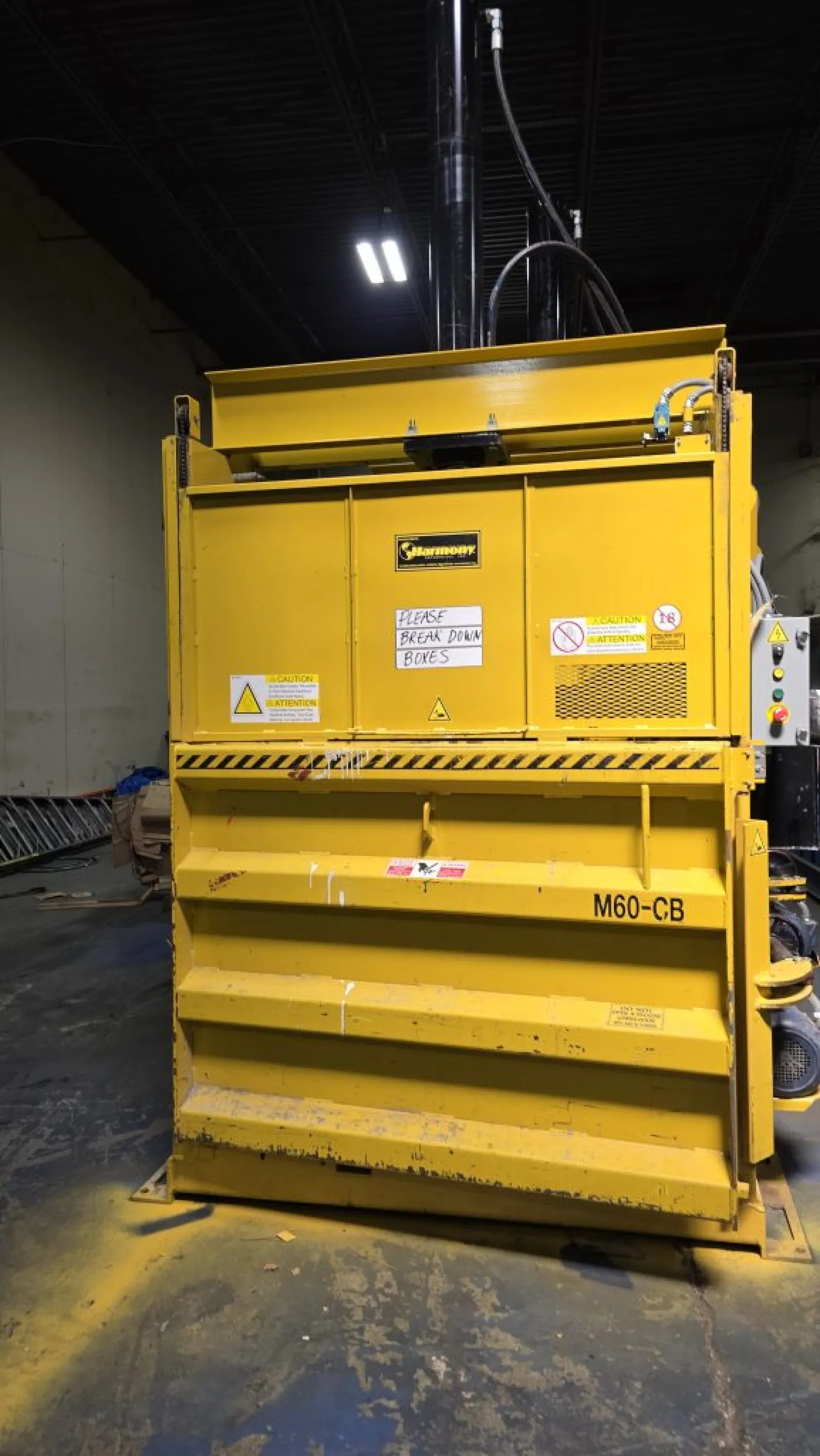Large yellow industrial baler machine labeled M60-CB in a warehouse with caution signs and instructions.