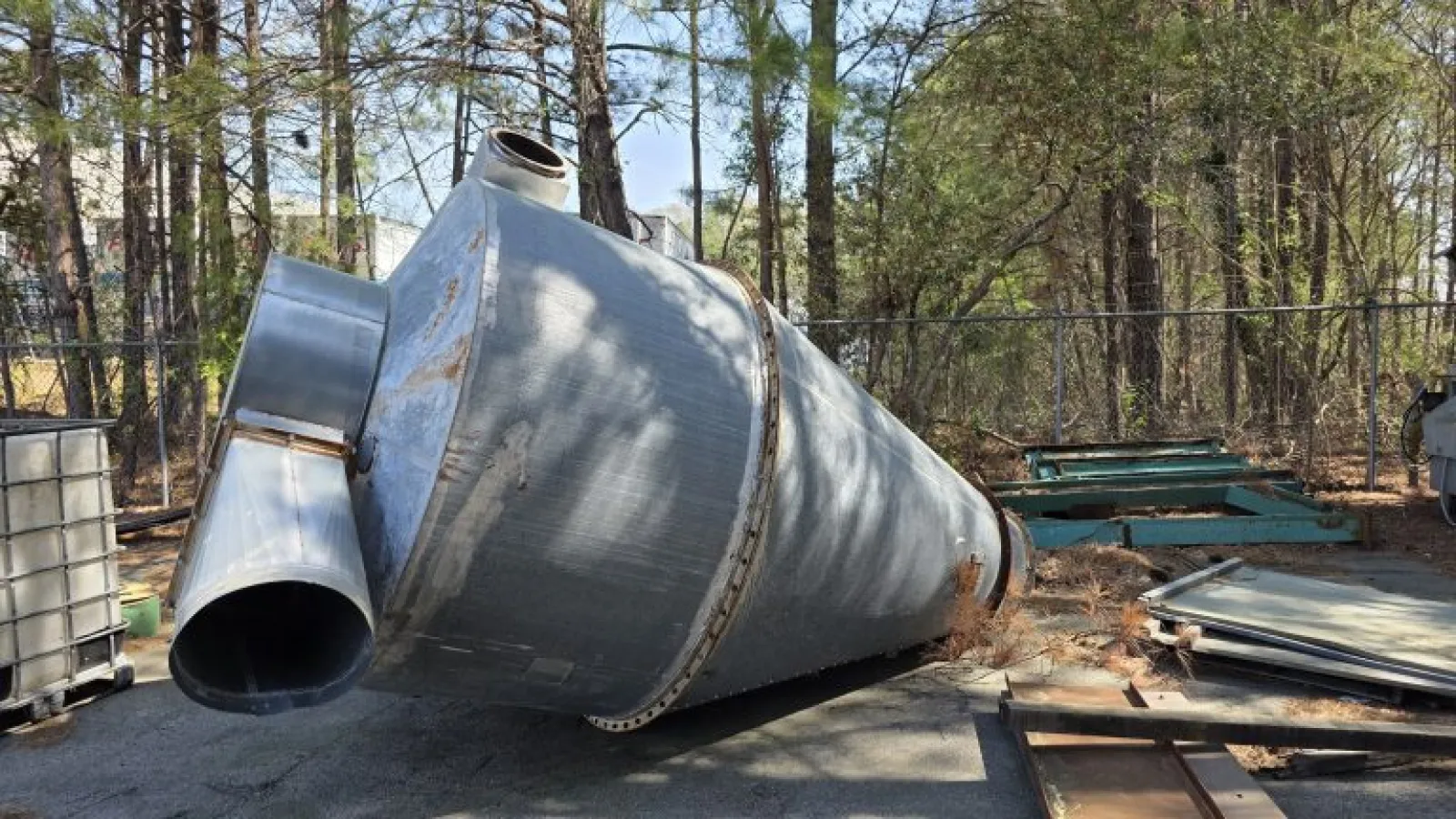 Large metal industrial silo lying on its side in an outdoor storage area surrounded by trees and equipment.