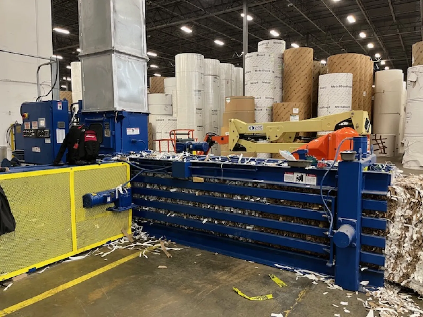 Industrial paper baler and large rolls of paper stacked in a warehouse with equipment and safety barriers.