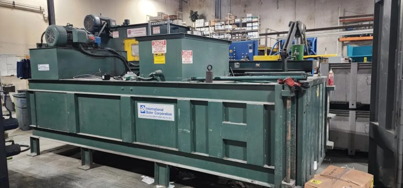 Large green industrial baler machine inside a warehouse with warning labels and company signage.