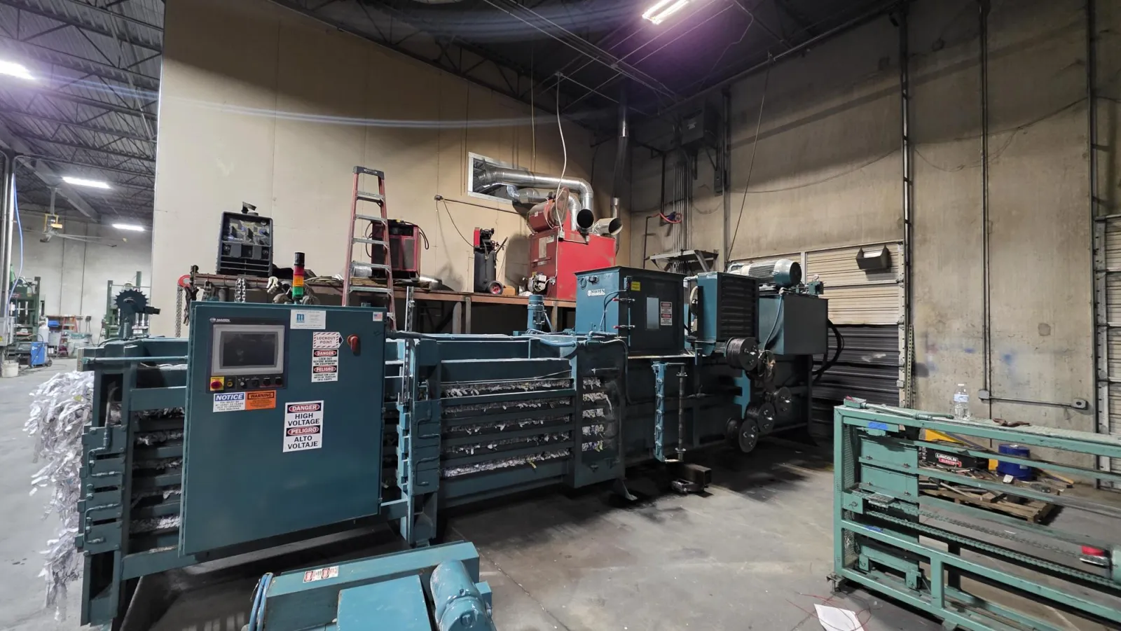 Large industrial paper baler machine compacting shredded paper in a spacious warehouse with equipment and ladder.