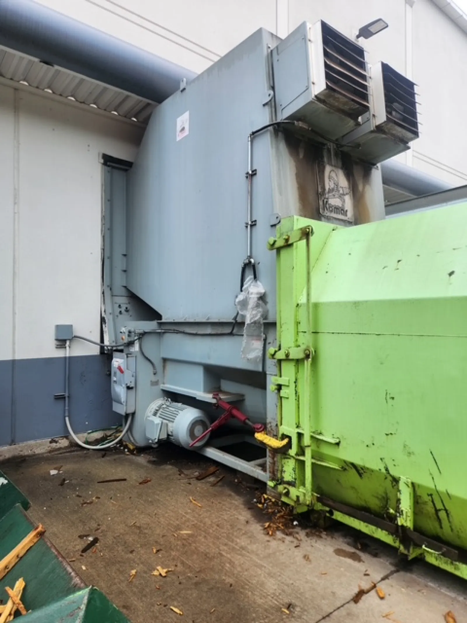 Industrial waste compactor with green container in a facility's outdoor area near concrete ground.