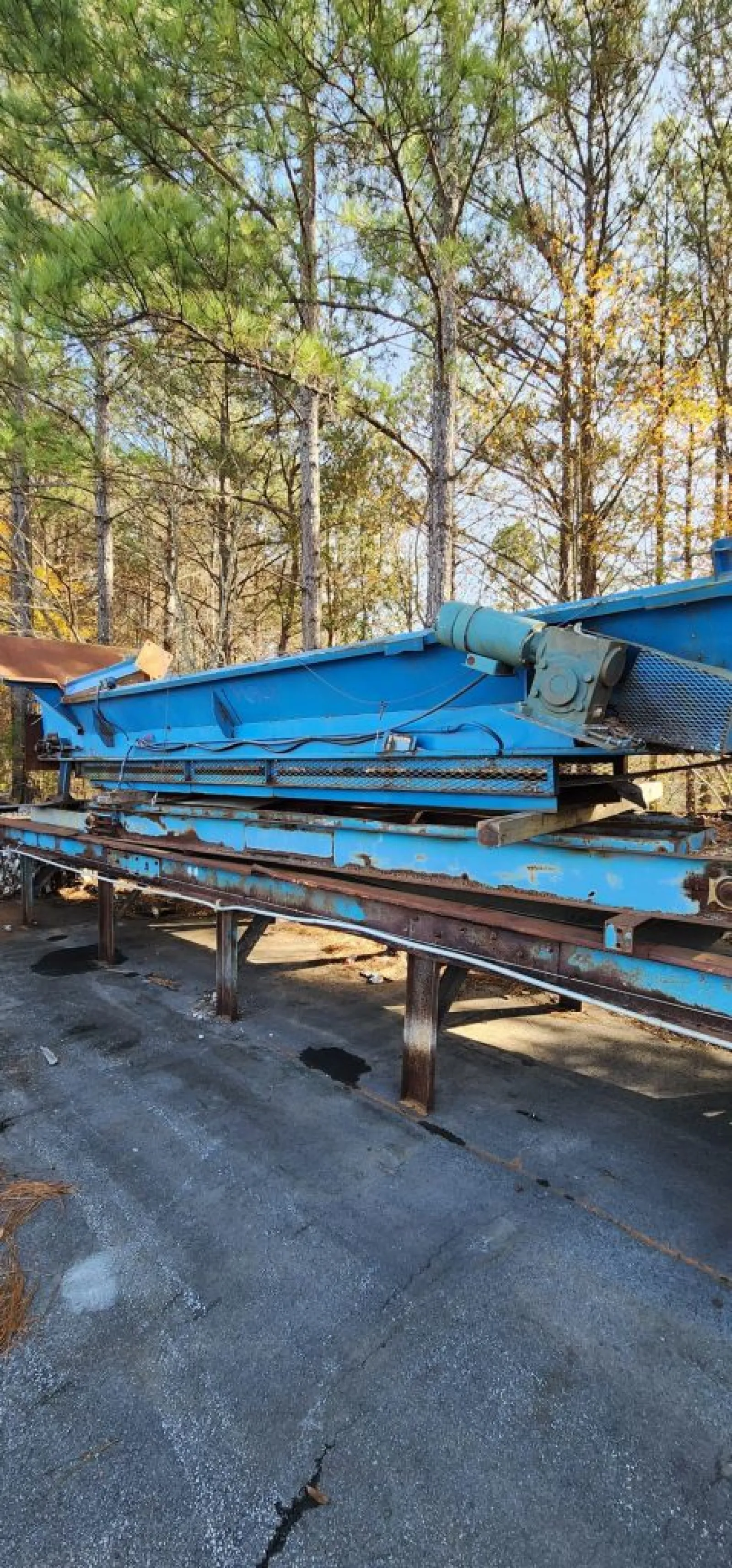 Large blue industrial conveyor belt machine outdoors near trees on cracked asphalt surface.