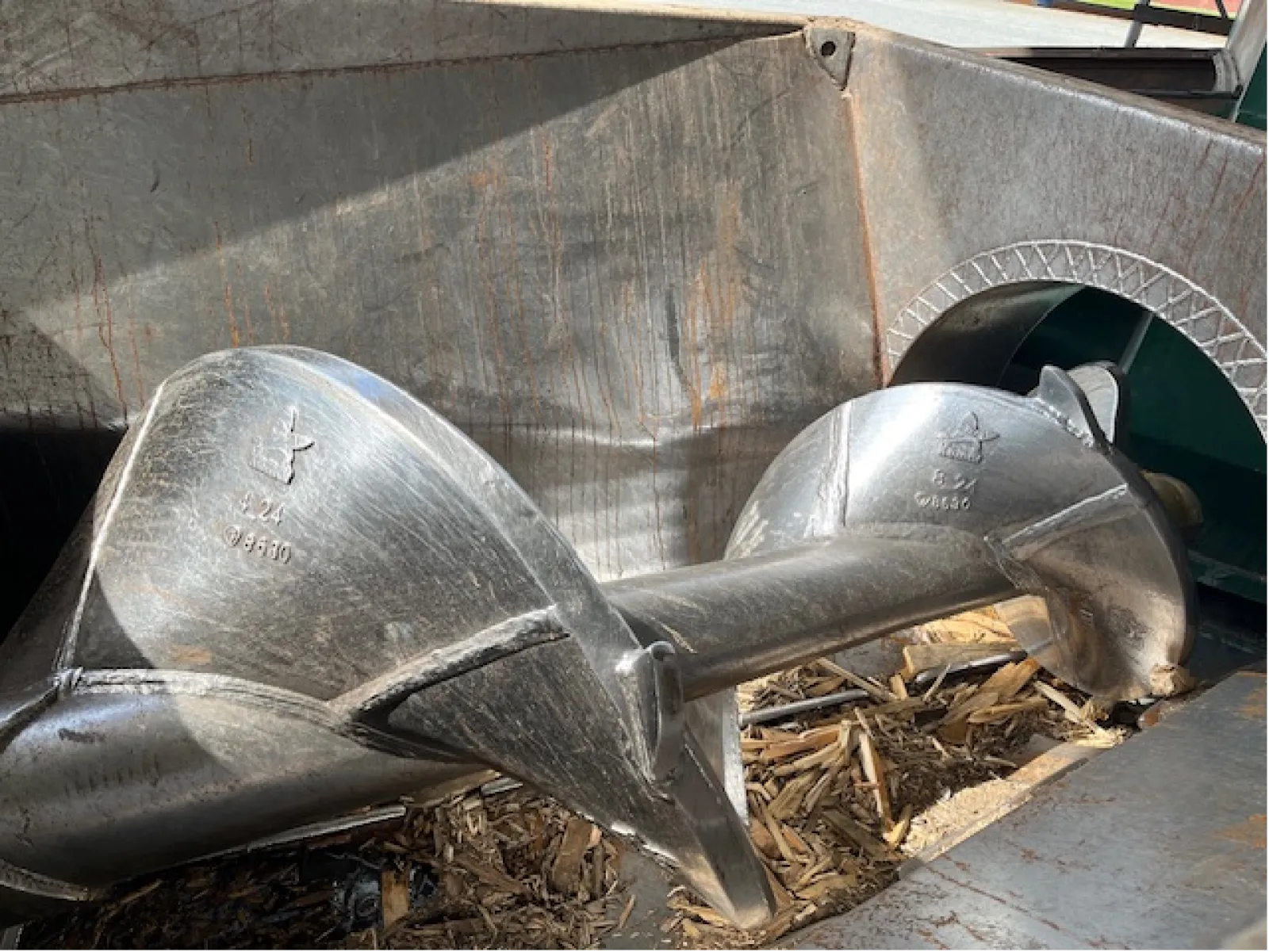 Close-up of a large industrial metal screw conveyor inside a container with wood chips.