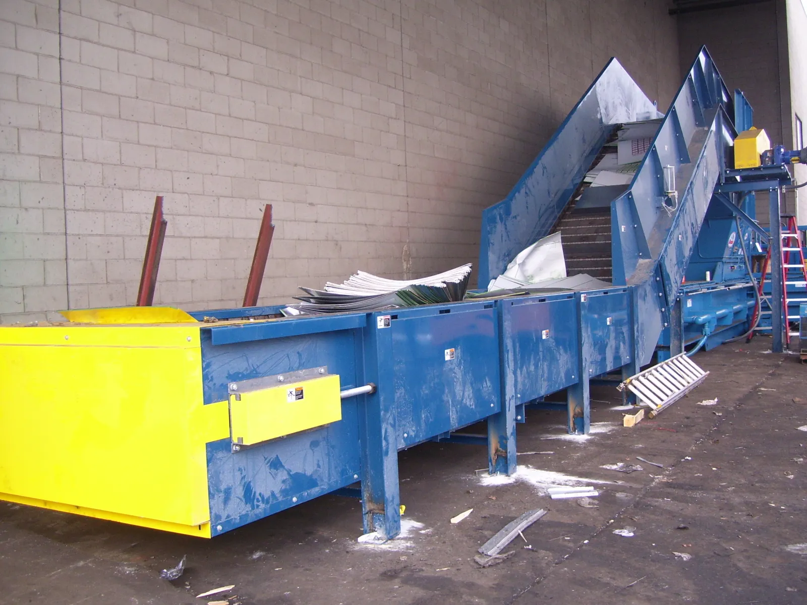 Industrial blue and yellow conveyor belt system inside warehouse with scattered debris on floor.
