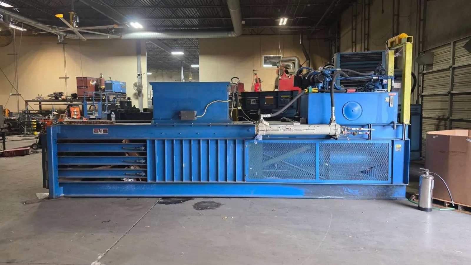 Large blue industrial baler machine inside a warehouse with concrete floor and metal shelves nearby.