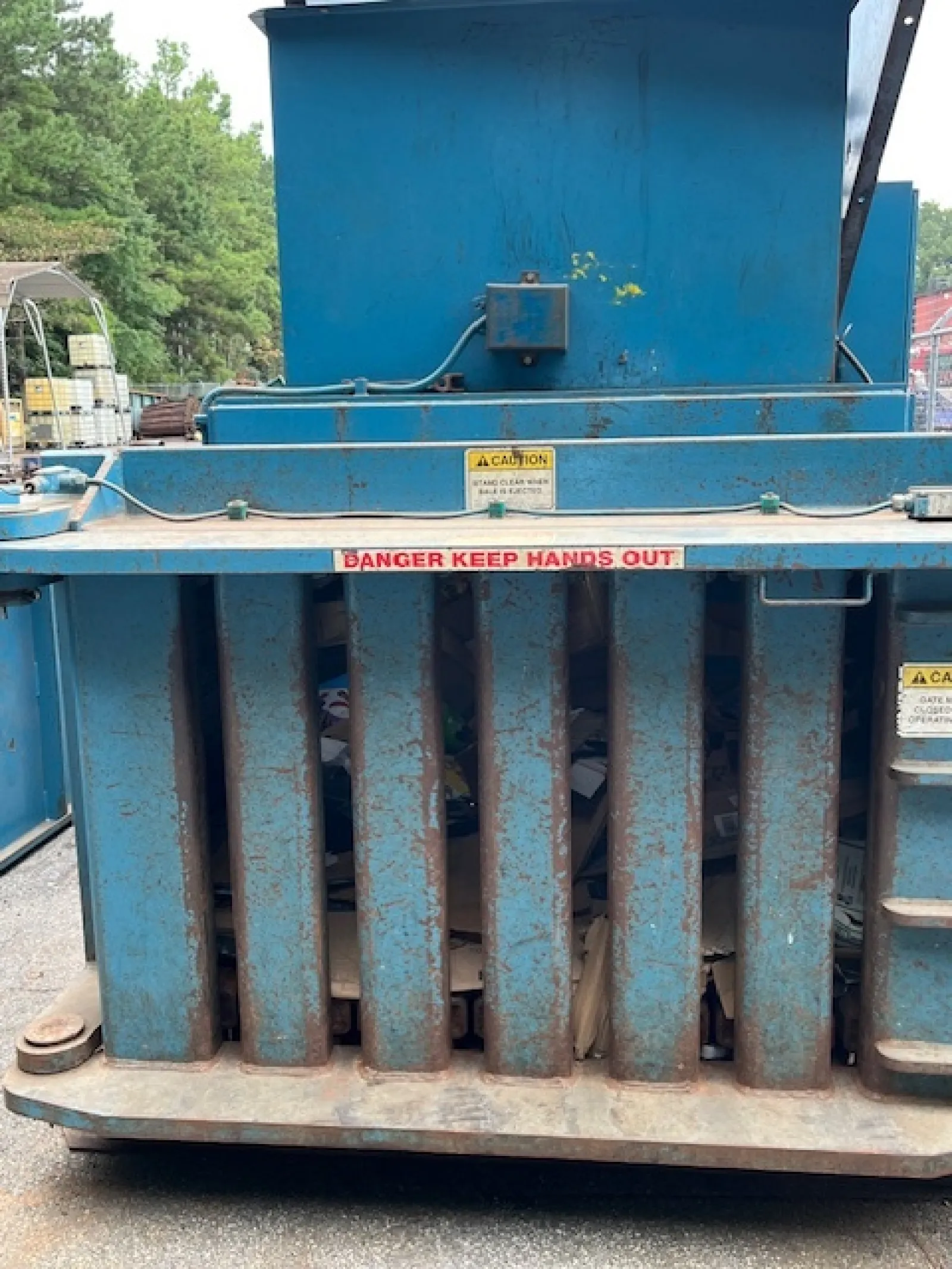 Rusty blue industrial trash compactor with caution signs and visible compressed waste inside.