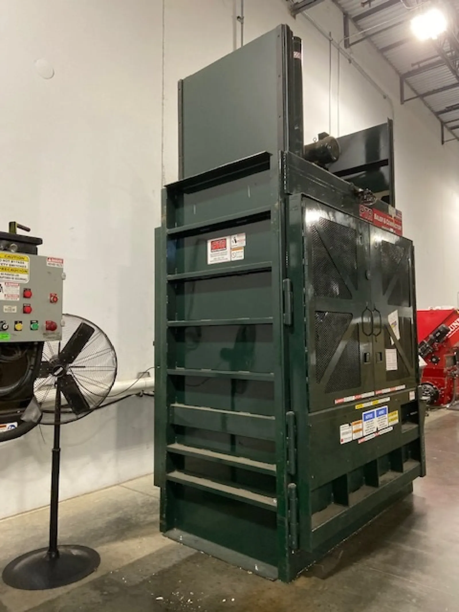 Large green industrial baler machine inside warehouse with control panel and standing fan nearby