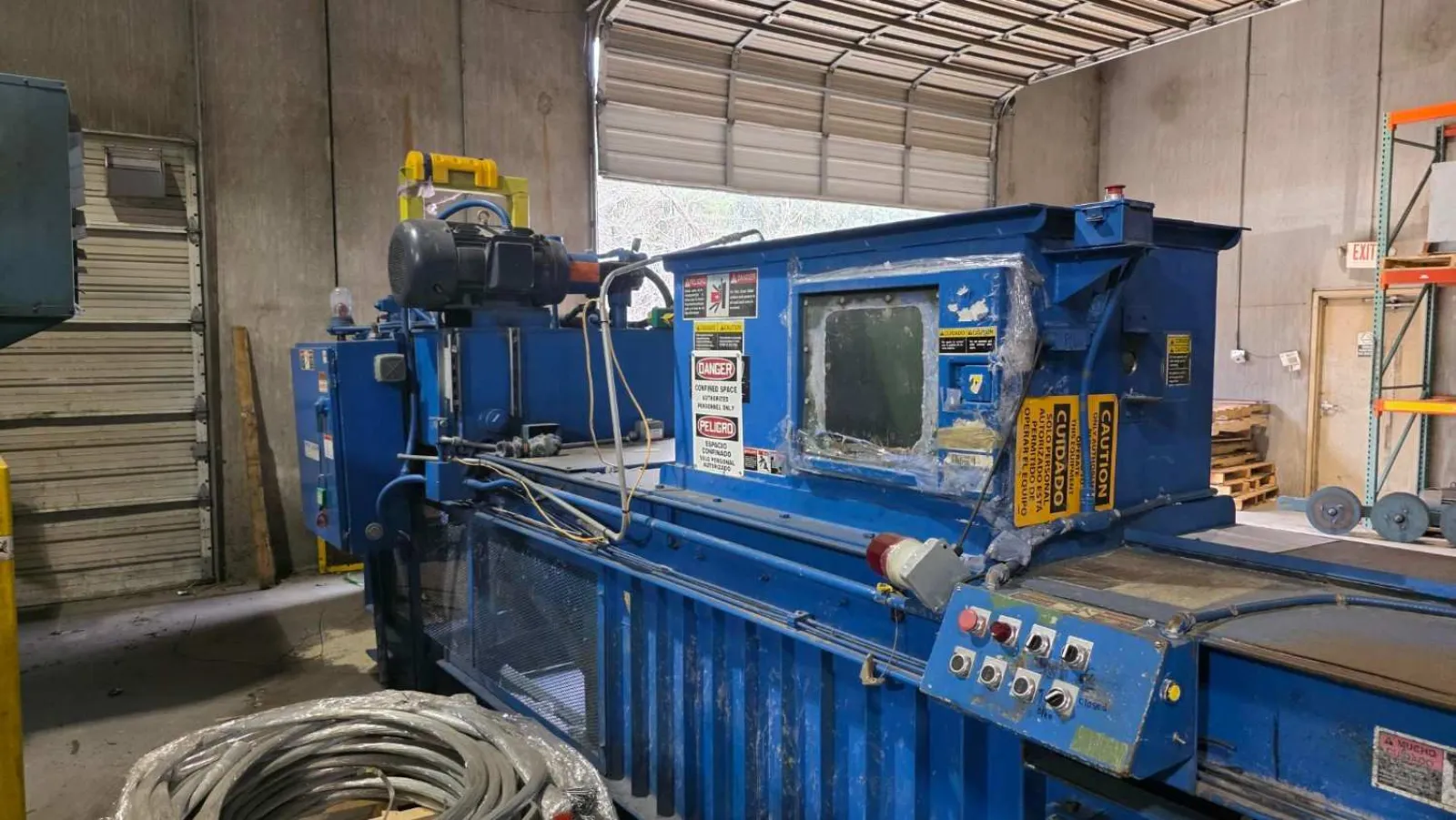 Blue industrial machine with control panel inside a warehouse near coiled cables and open garage door.