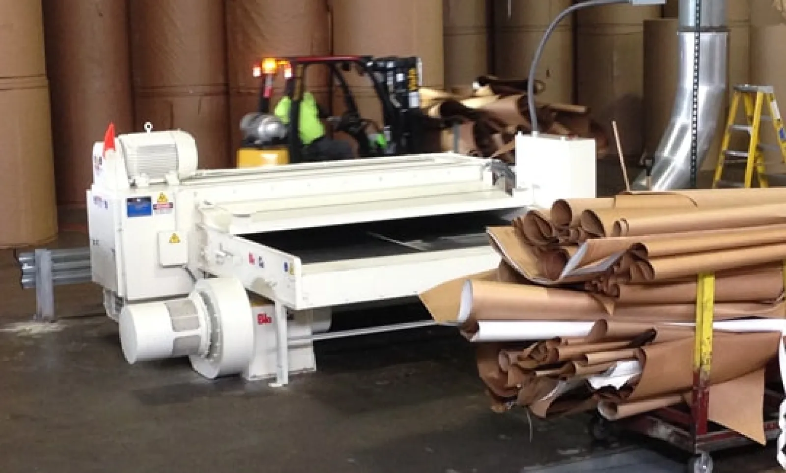 Industrial paper manufacturing equipment with large rolls of brown paper and forklift in warehouse.