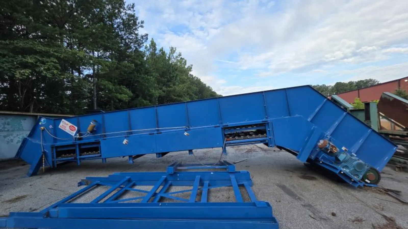 Large blue industrial conveyor belt system outdoors on gravel near trees and buildings under partly cloudy sky.