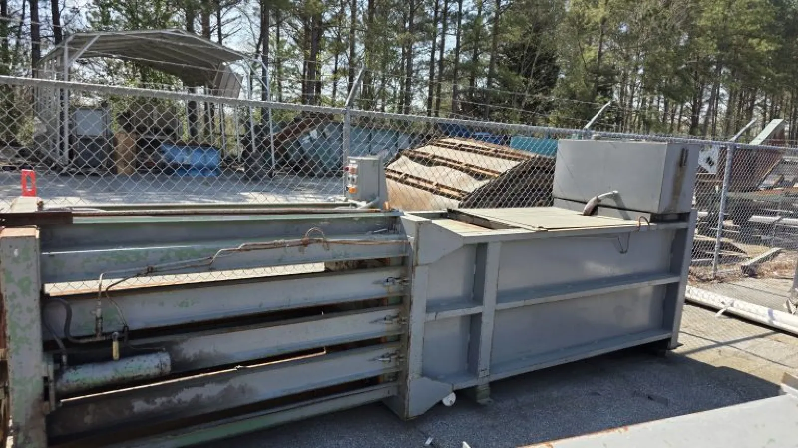 Large industrial metal baler machine for compacting recyclable materials outdoors in a fenced yard.