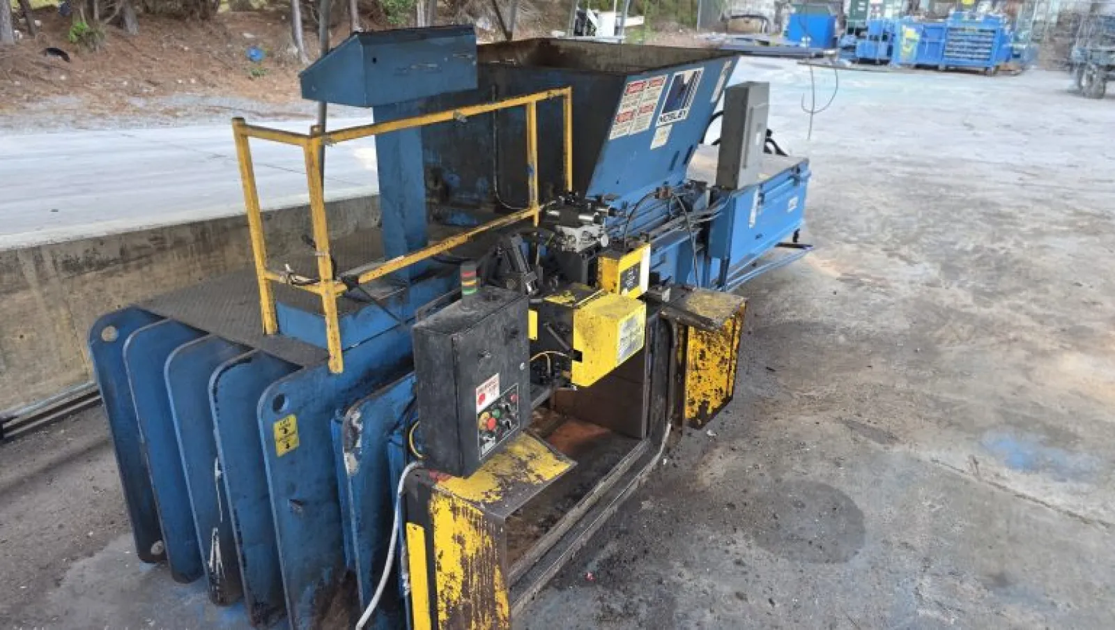 Industrial metal baler machine with blue and yellow parts in a warehouse setting on concrete floor.