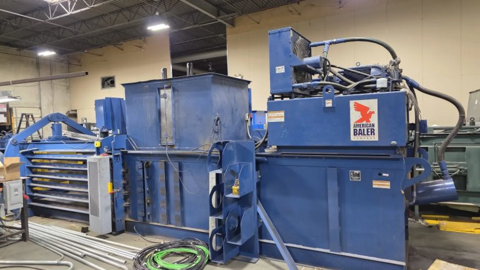 Industrial blue American Baler machine inside a warehouse with hydraulic components and cables on the floor.