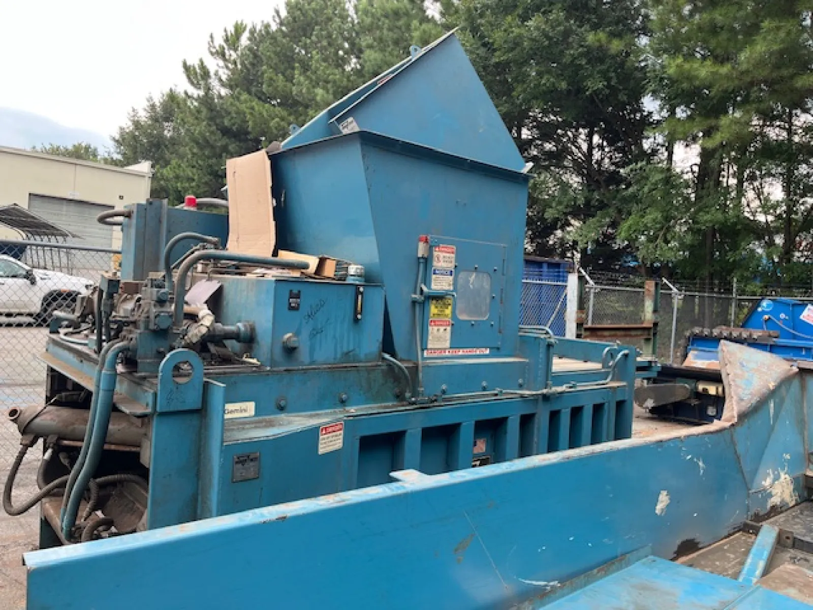 Large blue industrial machine with hydraulic components and chute in outdoor fenced area near trees and vehicles.