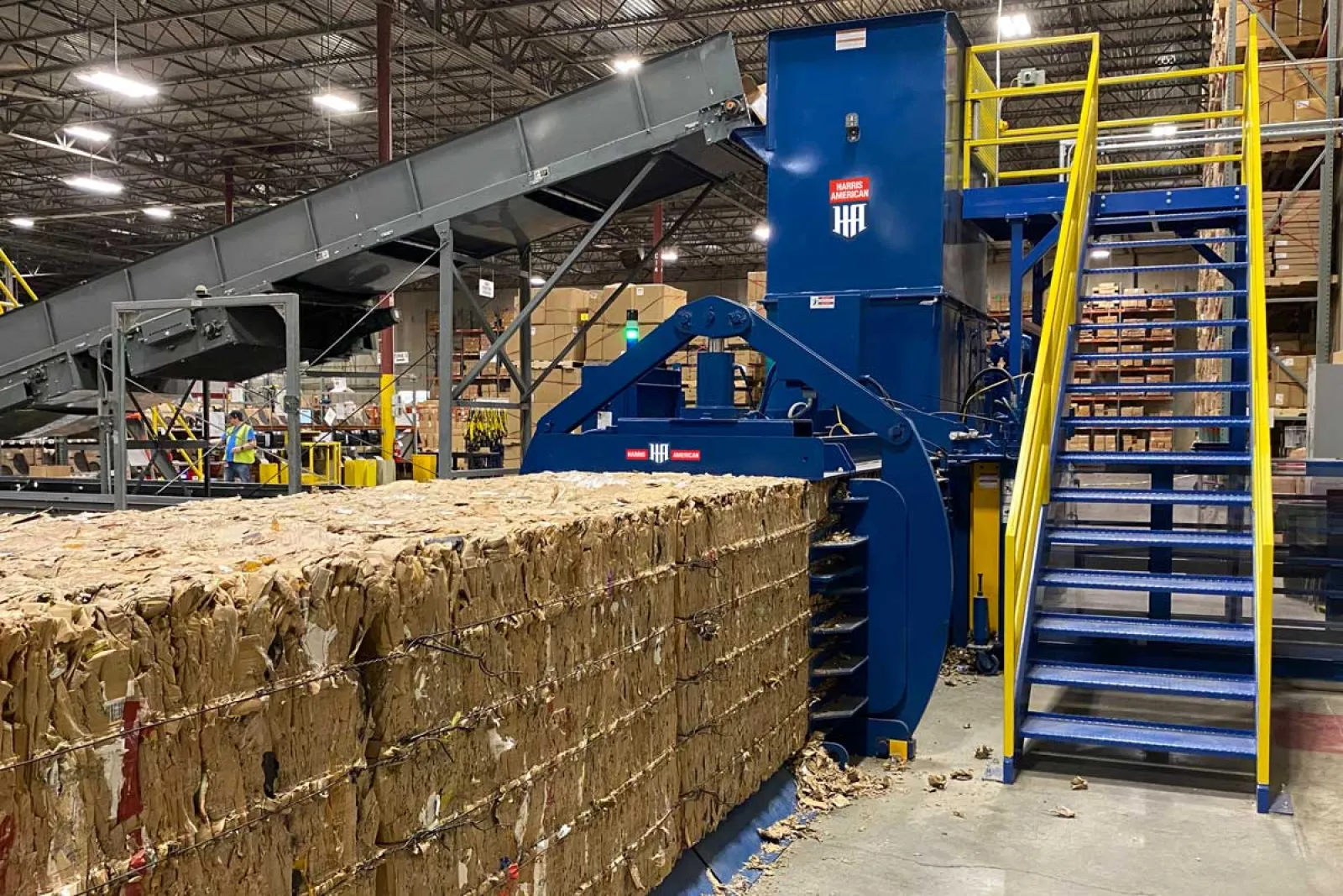 Large compressed bale of cardboard in an industrial recycling facility with machinery and workers in background