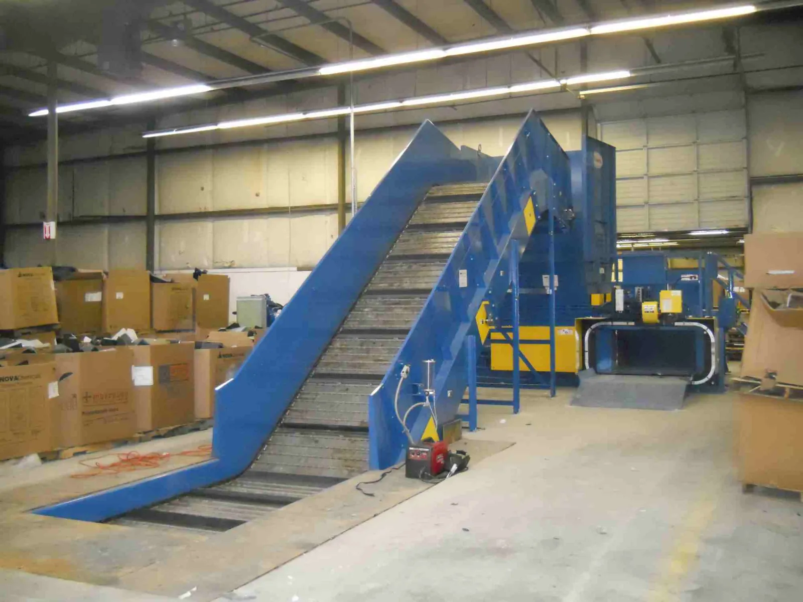 Blue industrial conveyor belt system in warehouse surrounded by cardboard boxes and equipment