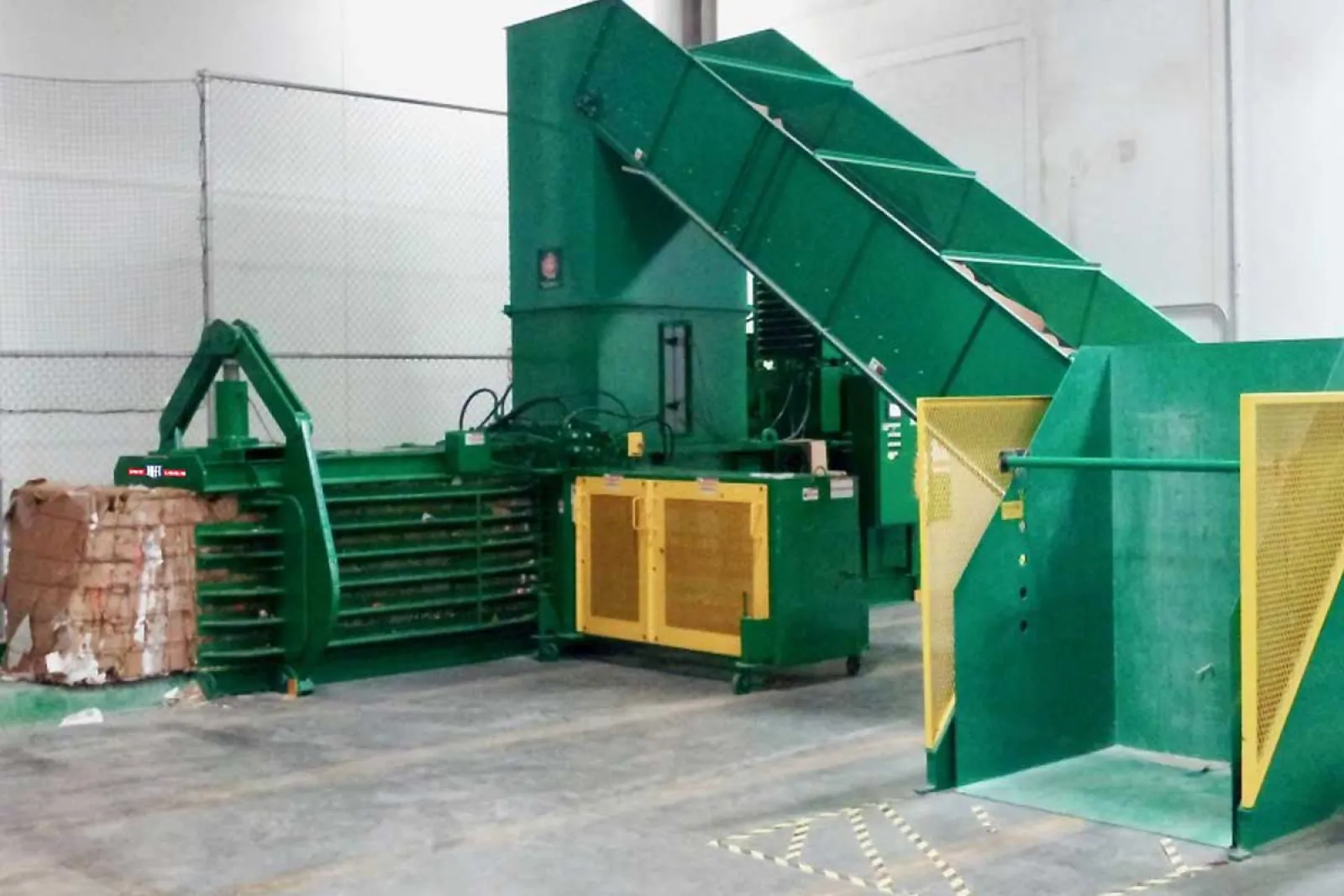 Large industrial green baler machine compressing cardboard waste in warehouse environment