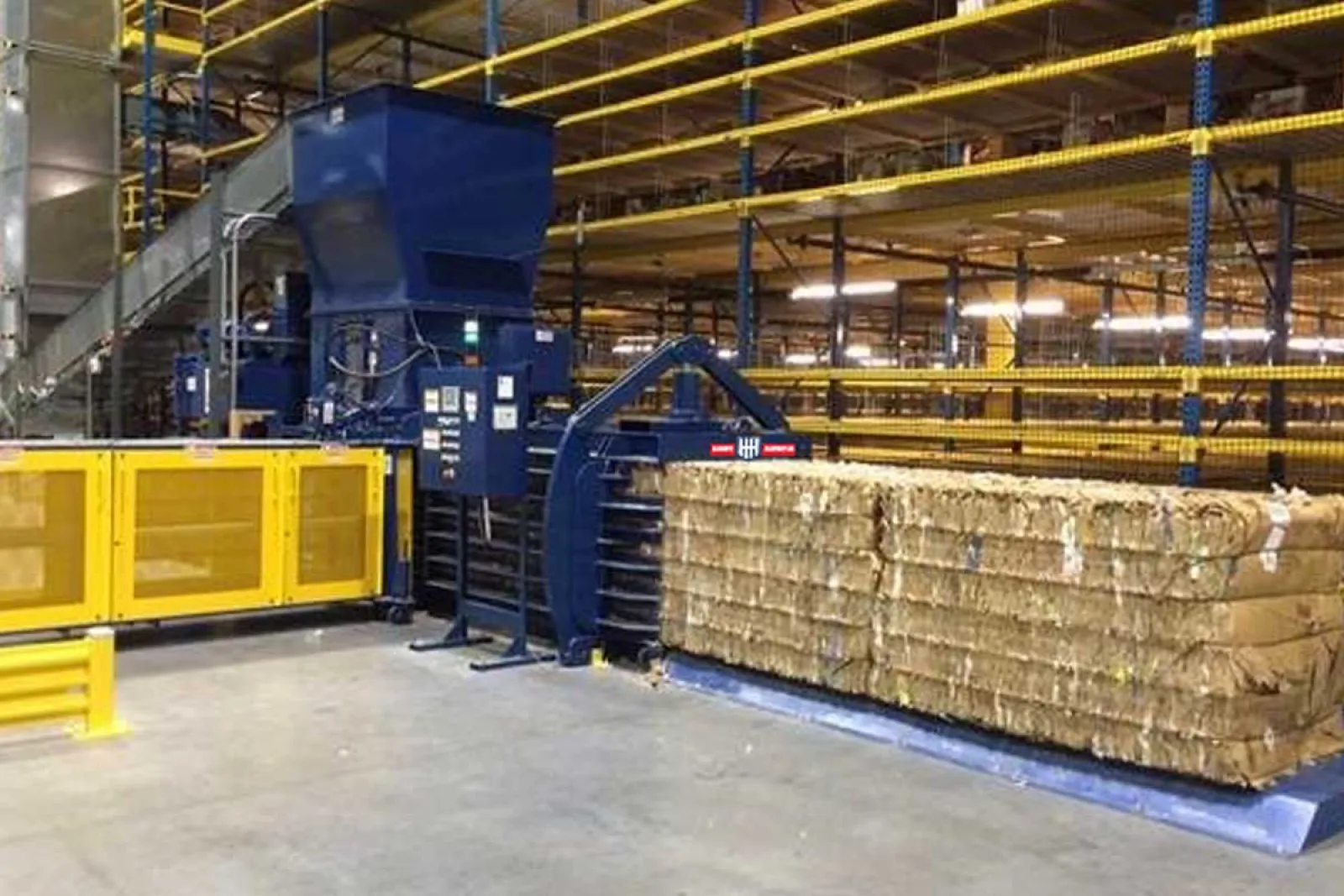 Automated industrial baler compressing cardboard bales in a warehouse with yellow safety barriers and metal racks.