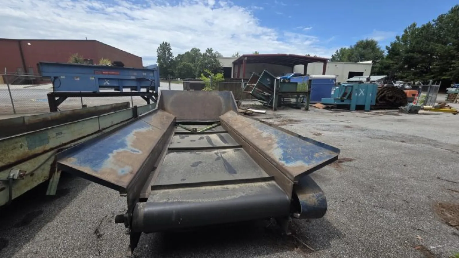 Industrial conveyor belt system outdoors with weathered metal components under a partly cloudy sky.