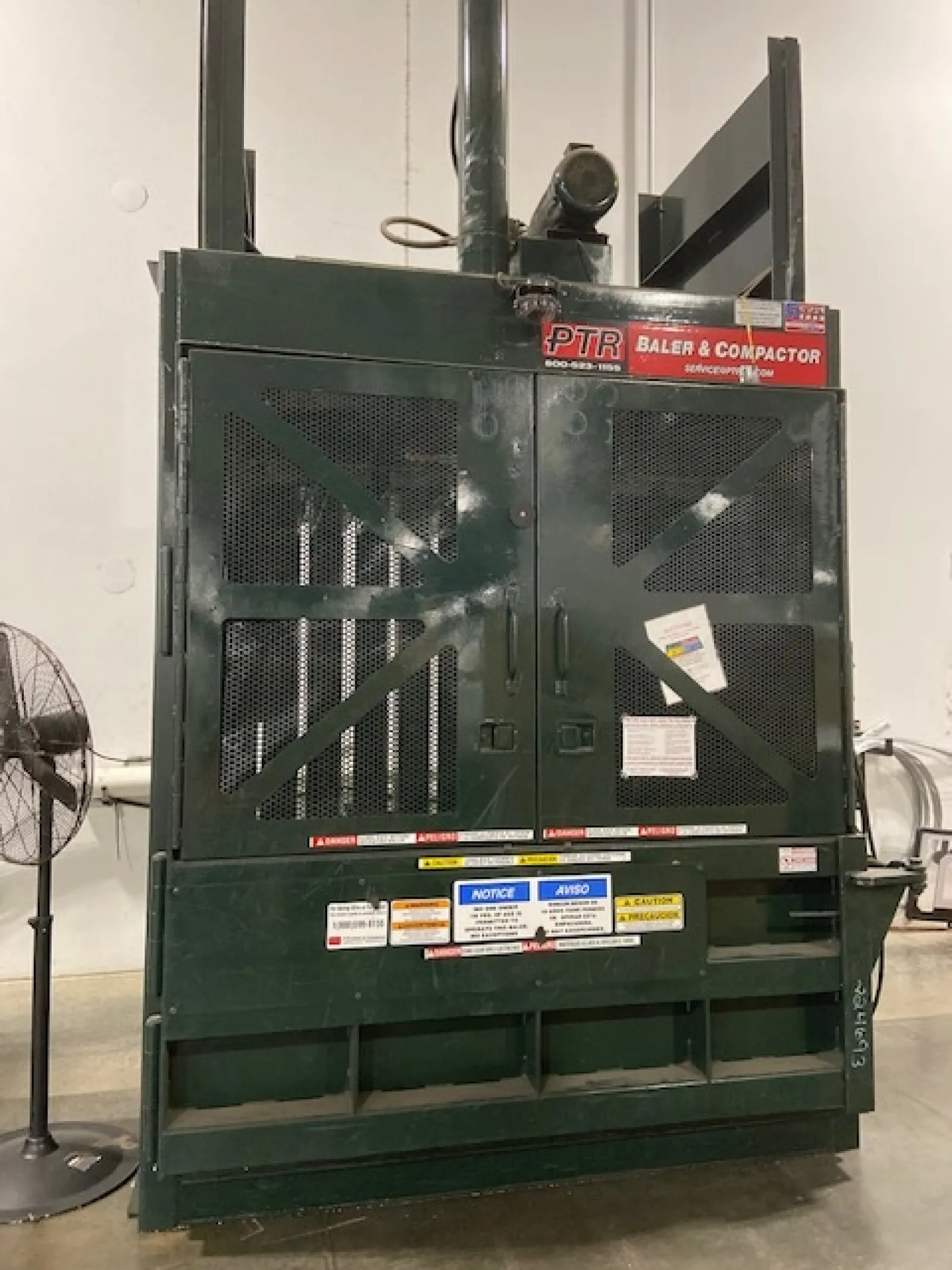 Industrial PTR baler and compactor machine in warehouse setting with caution labels and metal mesh doors.