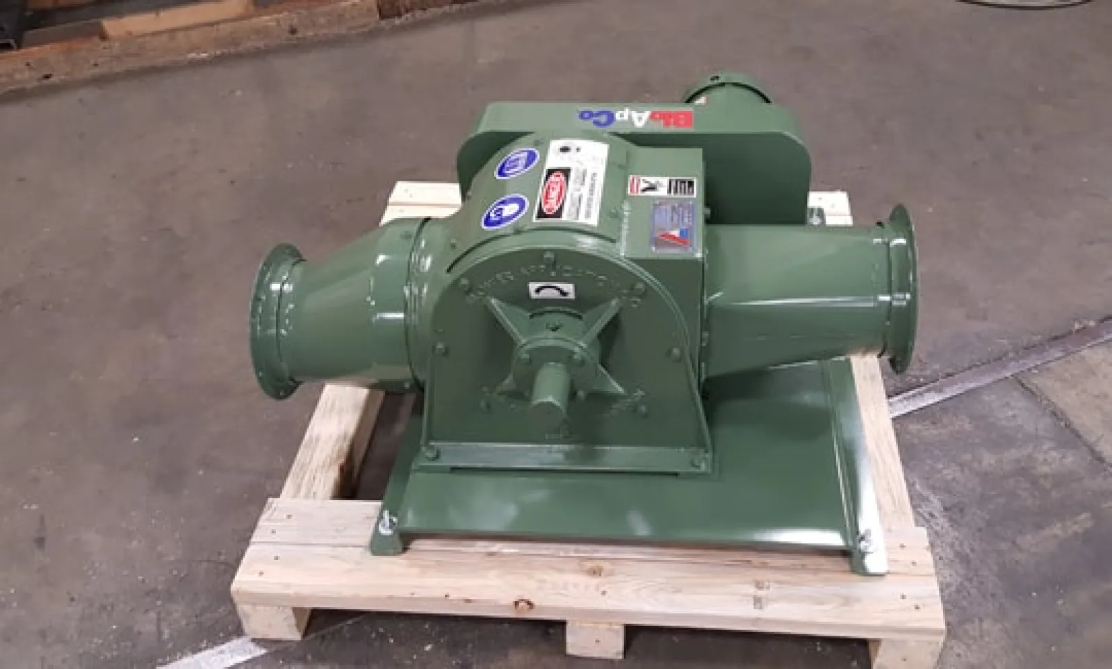 Green industrial blower mounted on a wooden pallet inside a workshop with concrete floor.