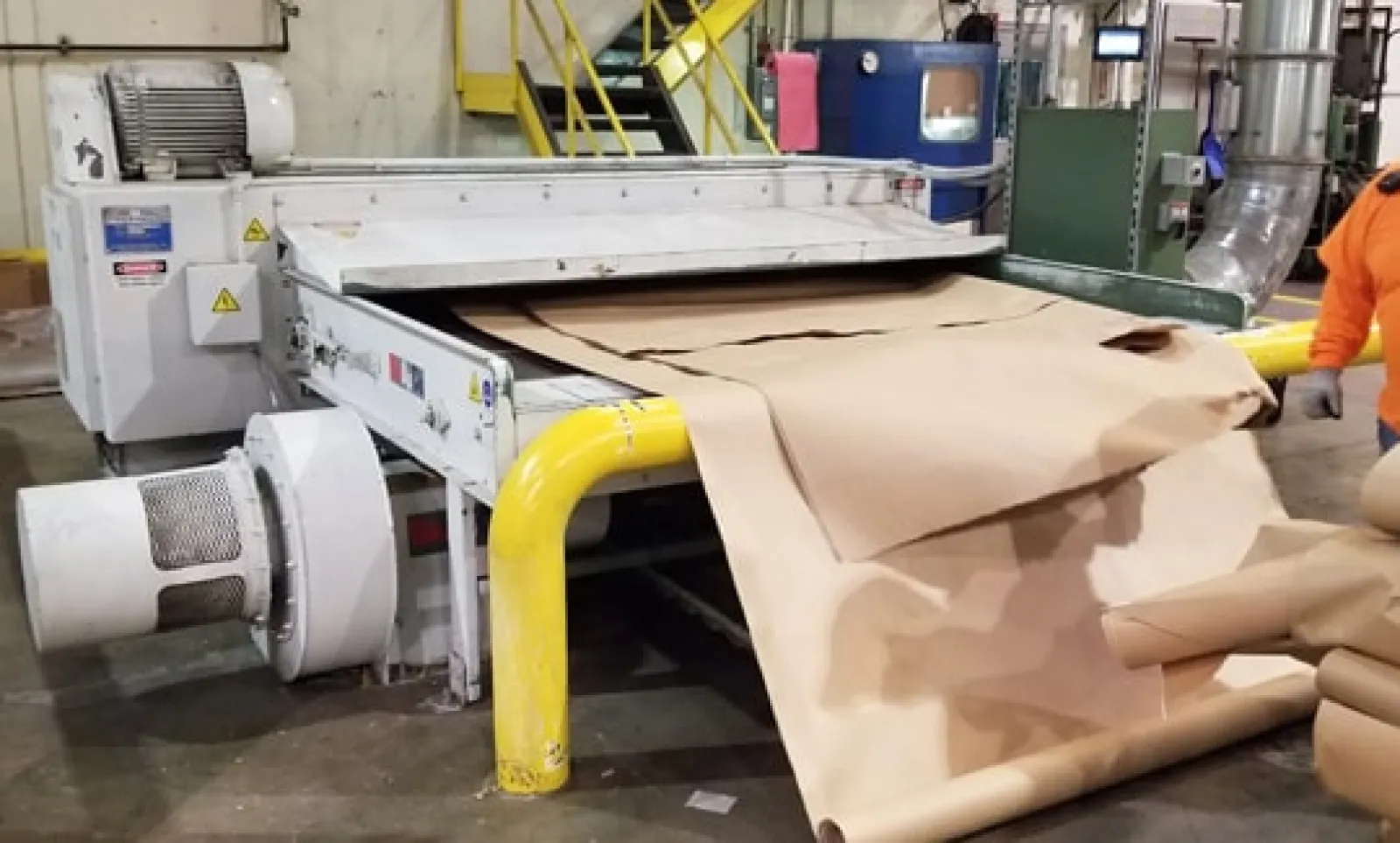 Industrial machine processing large rolls of brown kraft paper in a factory setting with yellow safety railings.