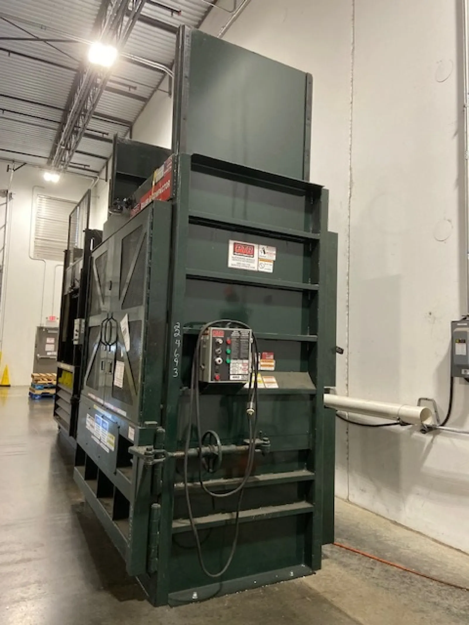 Large green industrial machine with control panel and cables in a warehouse with concrete floor and high ceiling.