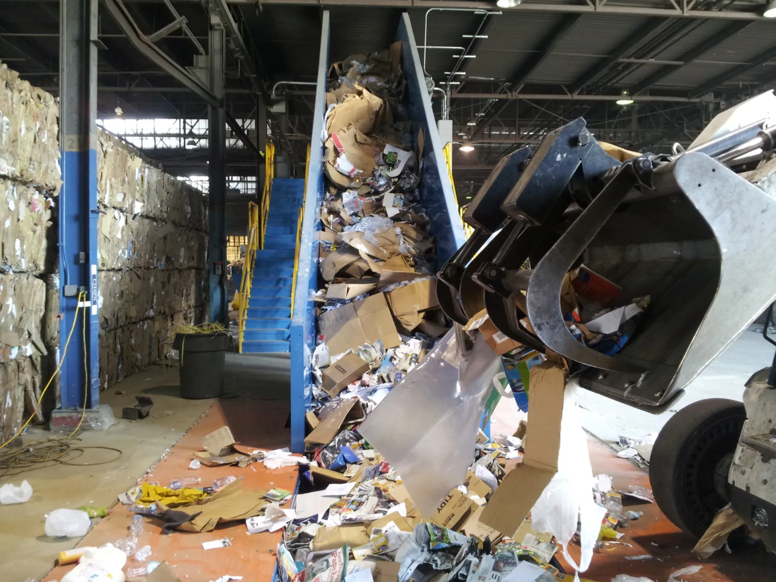 Recycling facility conveyor belt loaded with cardboard and paper waste being handled by industrial machinery.