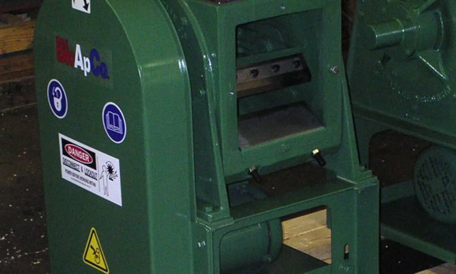 Green industrial machine with warning labels and safety signs in a workshop setting.