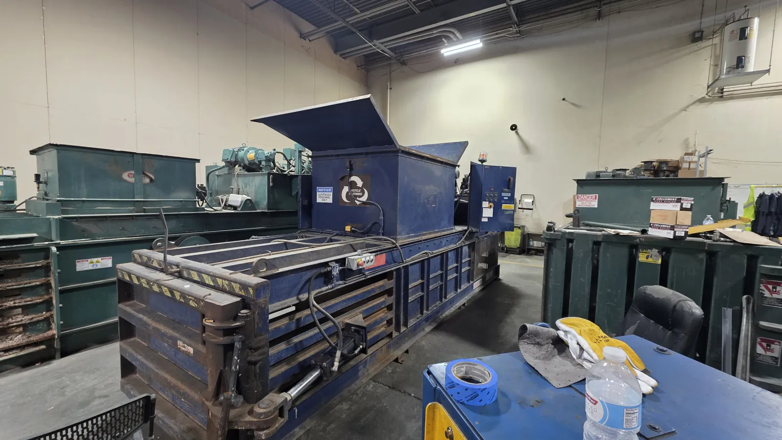 Industrial warehouse with large blue metal baler machine and nearby recycling bins in a sorting facility.