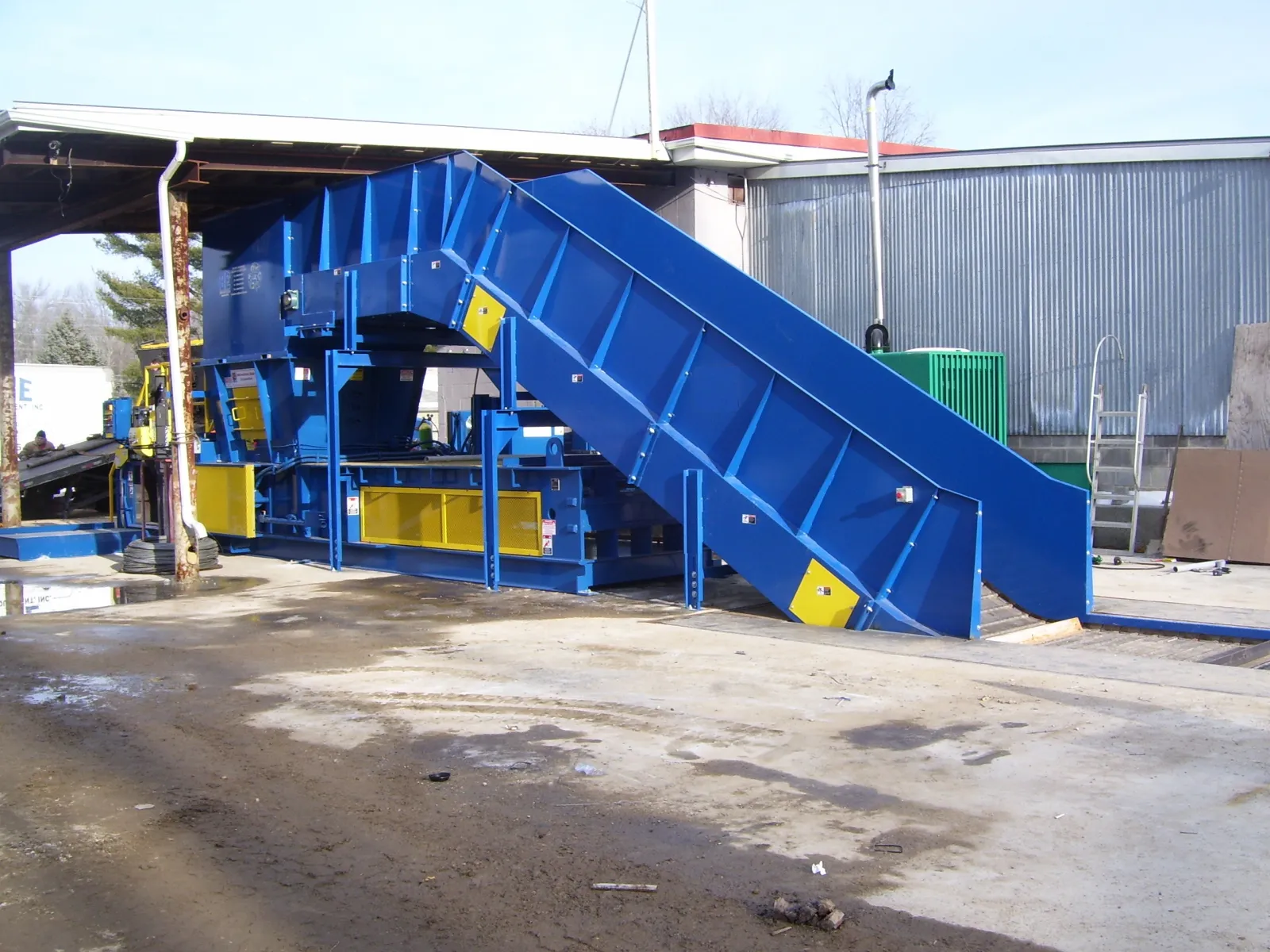 Large blue industrial machine with conveyor belt outdoors next to warehouse building on concrete ground.