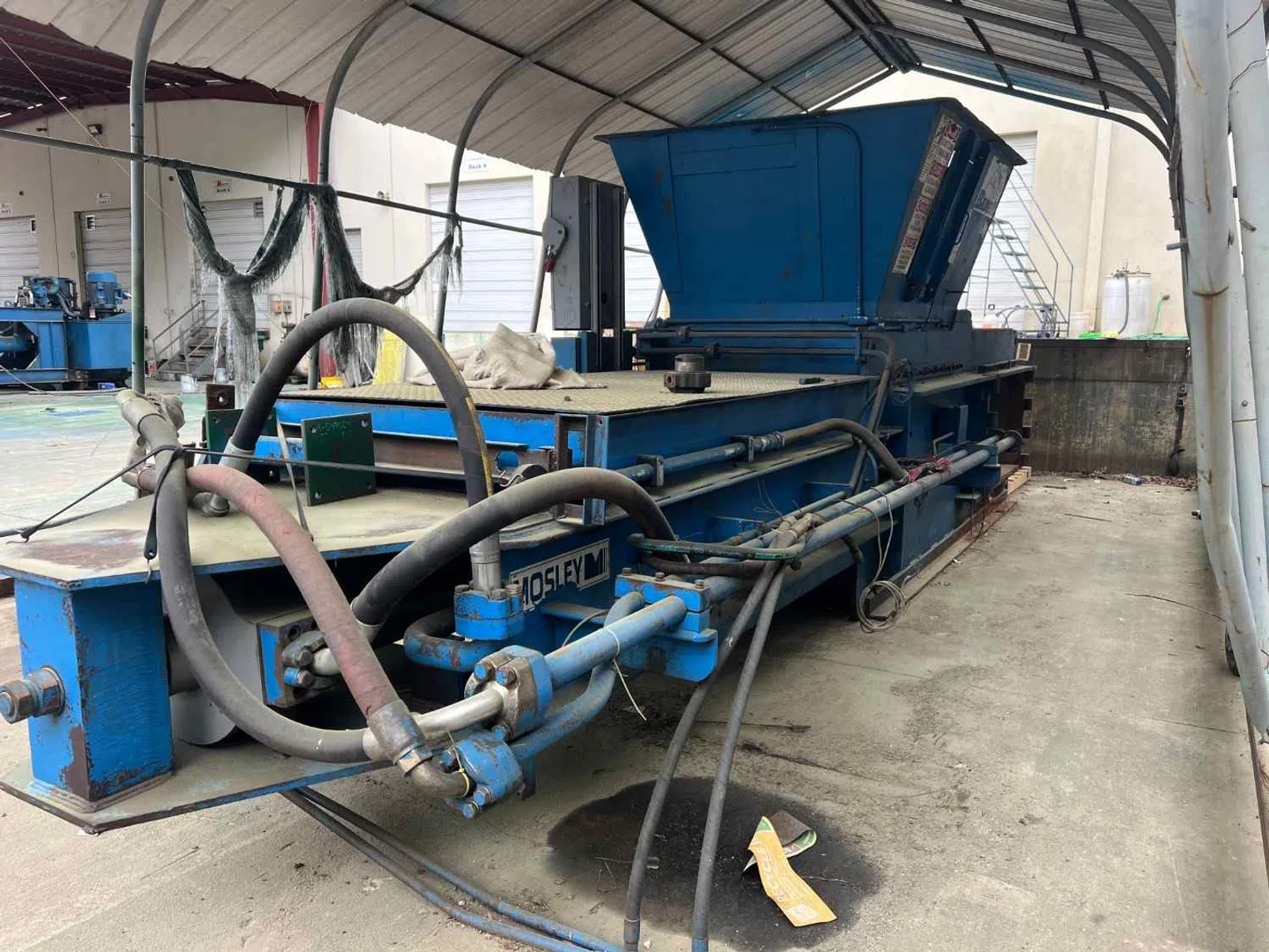 Large industrial blue metal machine with hoses inside a covered workspace on concrete floor.
