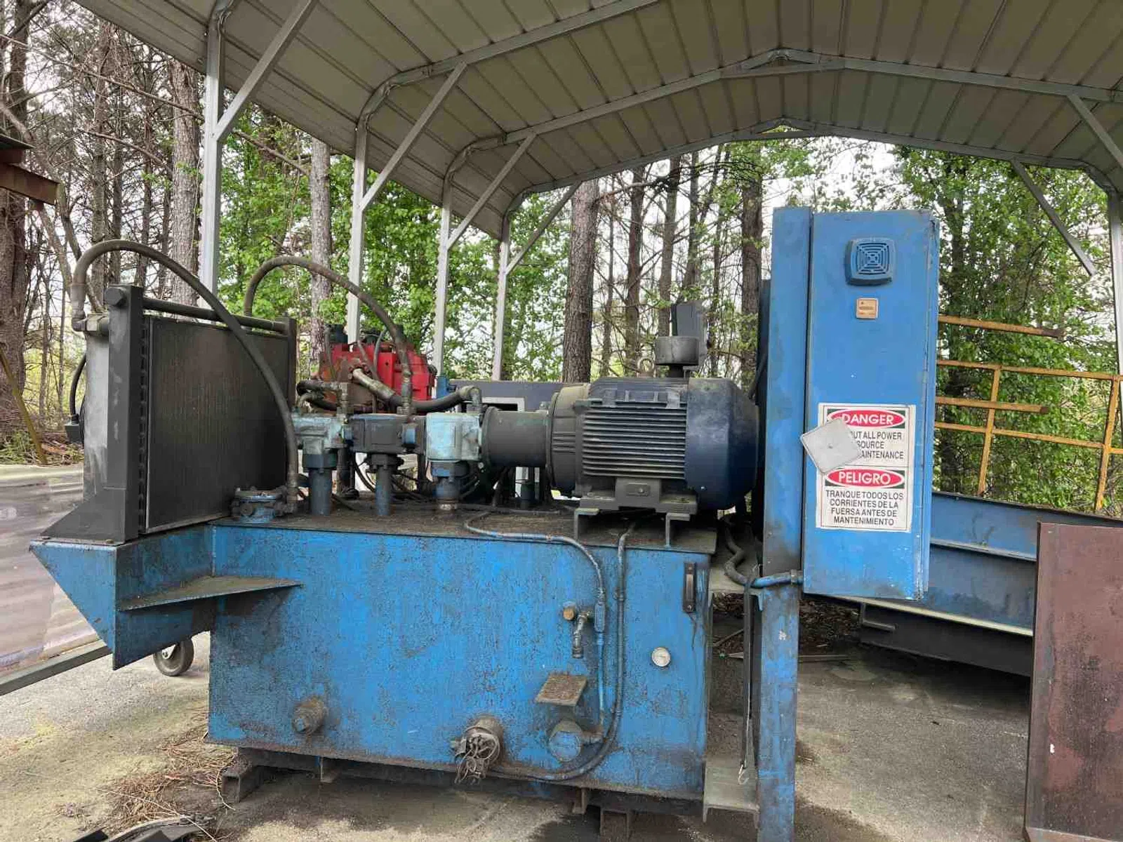 Industrial blue hydraulic machine with electric motor under a metal canopy outdoors surrounded by trees.
