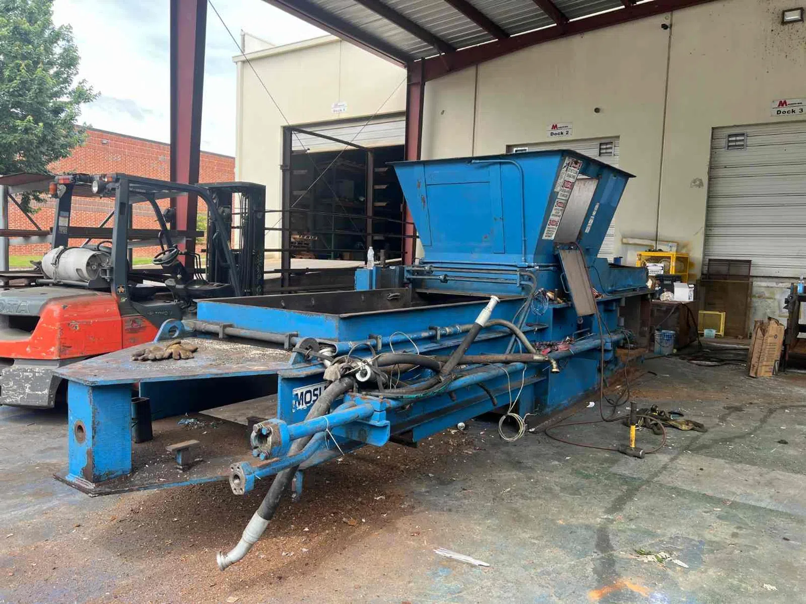 Industrial blue metal equipment with hoses and pipes inside warehouse near forklift and loading docks.
