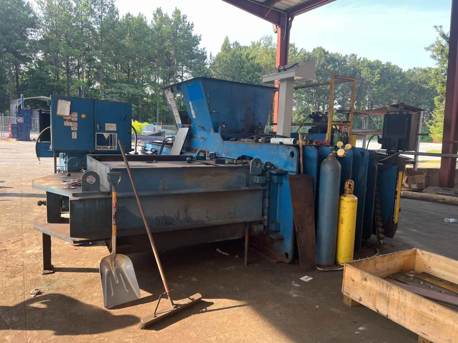 Industrial metal baler machine with gas cylinders under a metal roof in an outdoor workshop area.