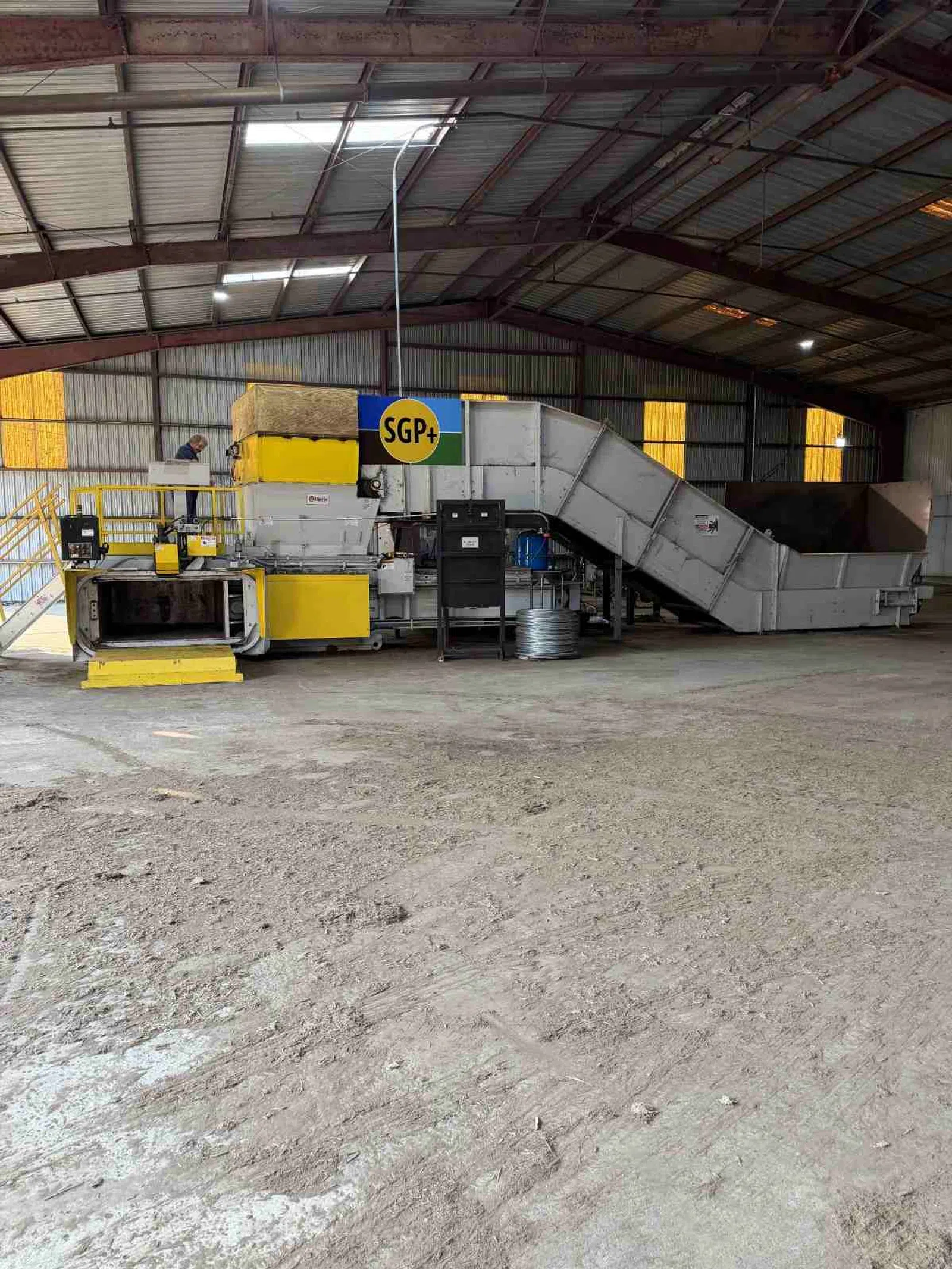 Industrial processing machine with yellow and grey parts inside a large warehouse with metal roof beams.