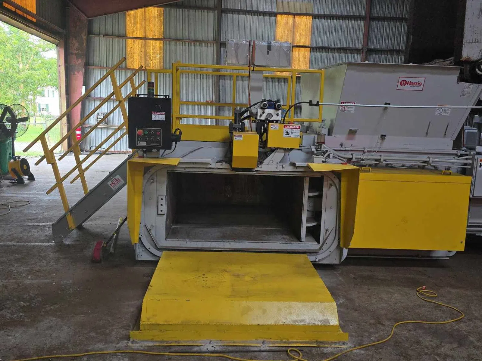 Industrial yellow waste compactor machine inside a warehouse with safety railings and control panel.