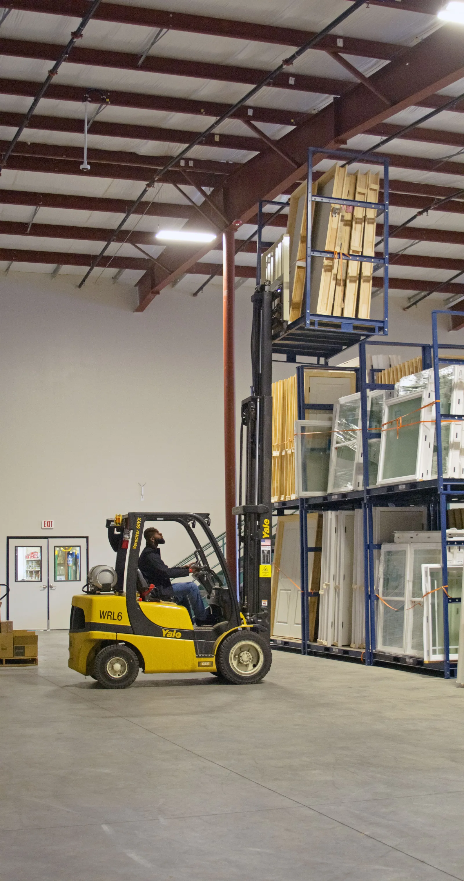 Man operating yellow Yale forklift lifting wooden panels in a spacious warehouse with organized shelves.