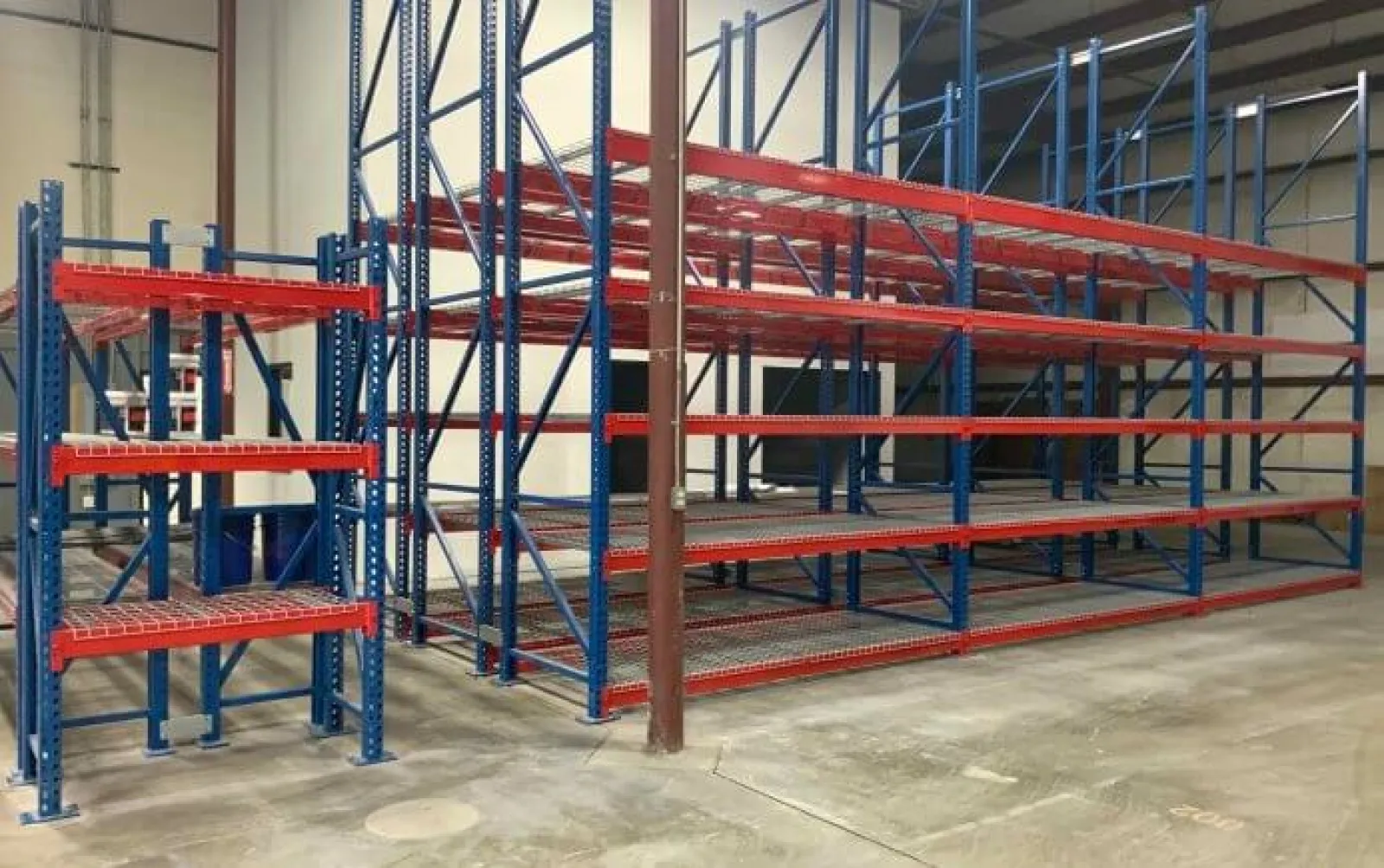 Empty industrial blue and red metal storage racks on a concrete floor inside a warehouse.