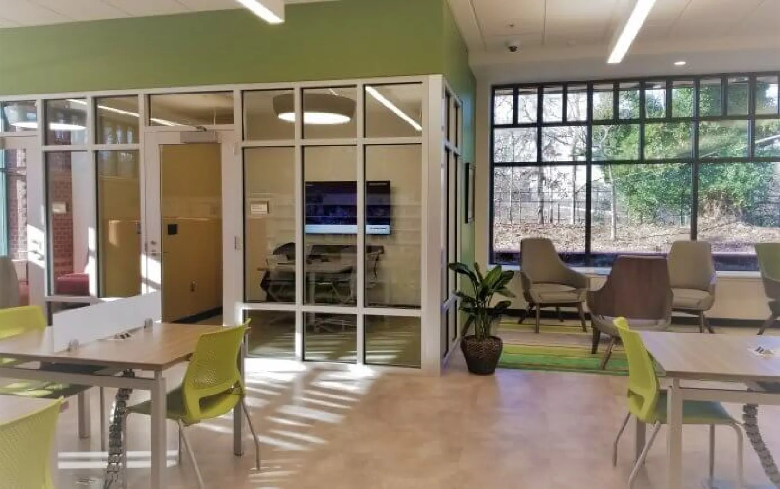 Modern office space with glass meeting room, green chairs, large window, and natural light filled room.