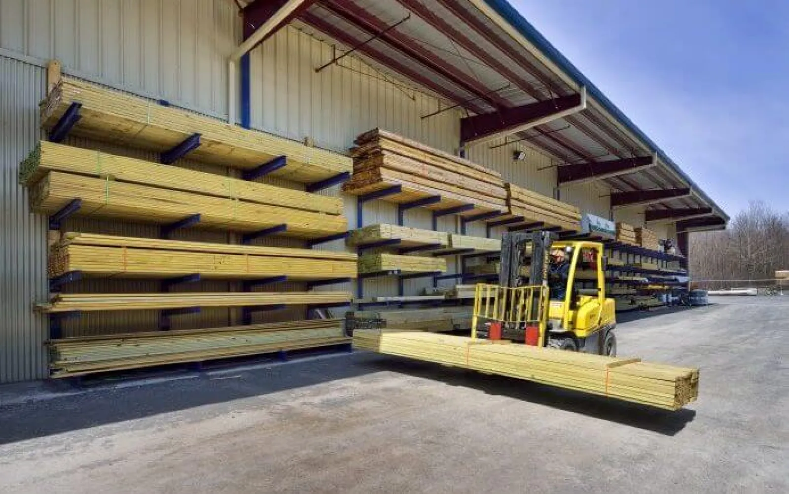 Yellow forklift carrying long wooden planks outside a lumber store with stacks of wood on metal racks under roof.