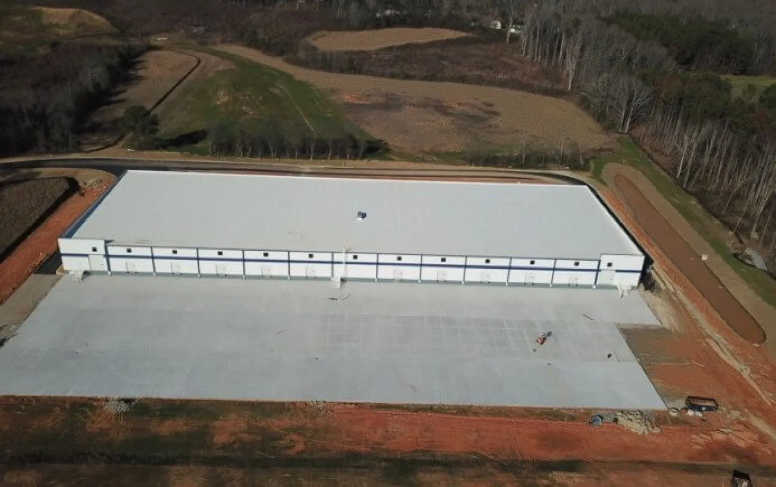 Aerial view of a large new industrial warehouse with a white roof surrounded by cleared land and forest.