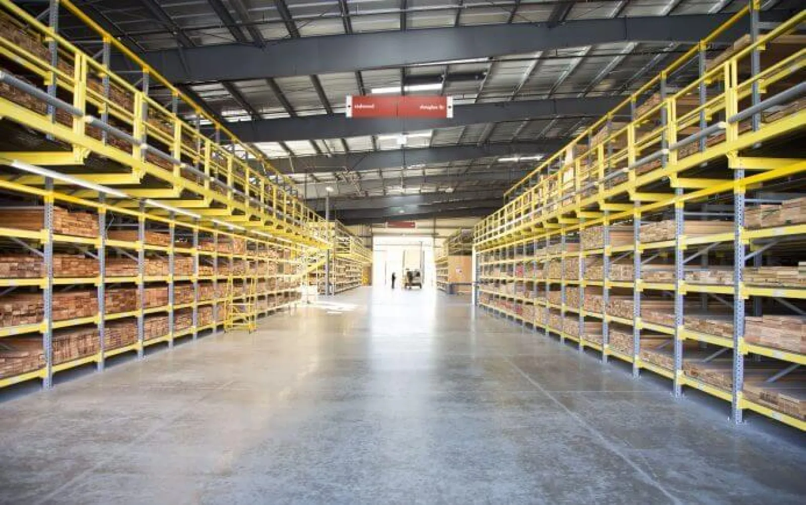 Spacious warehouse with yellow industrial shelving stocked with wooden planks under a metal roof with bright lighting.