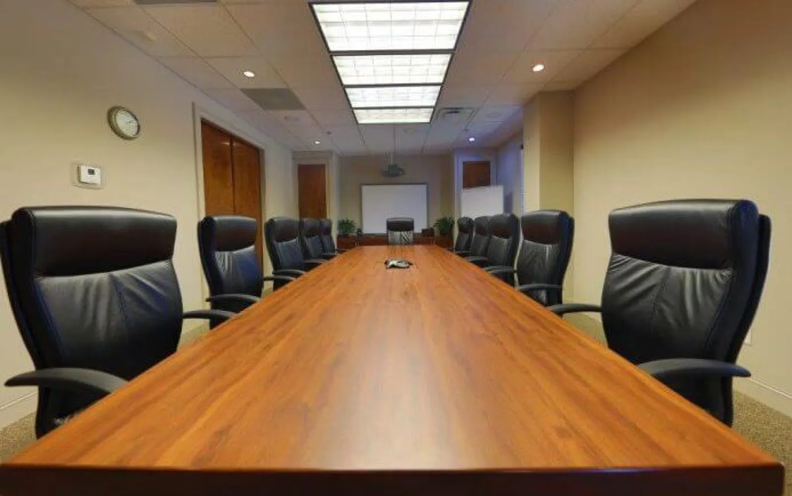 Spacious modern conference room with long wooden table, black leather chairs, projector, and neutral walls.