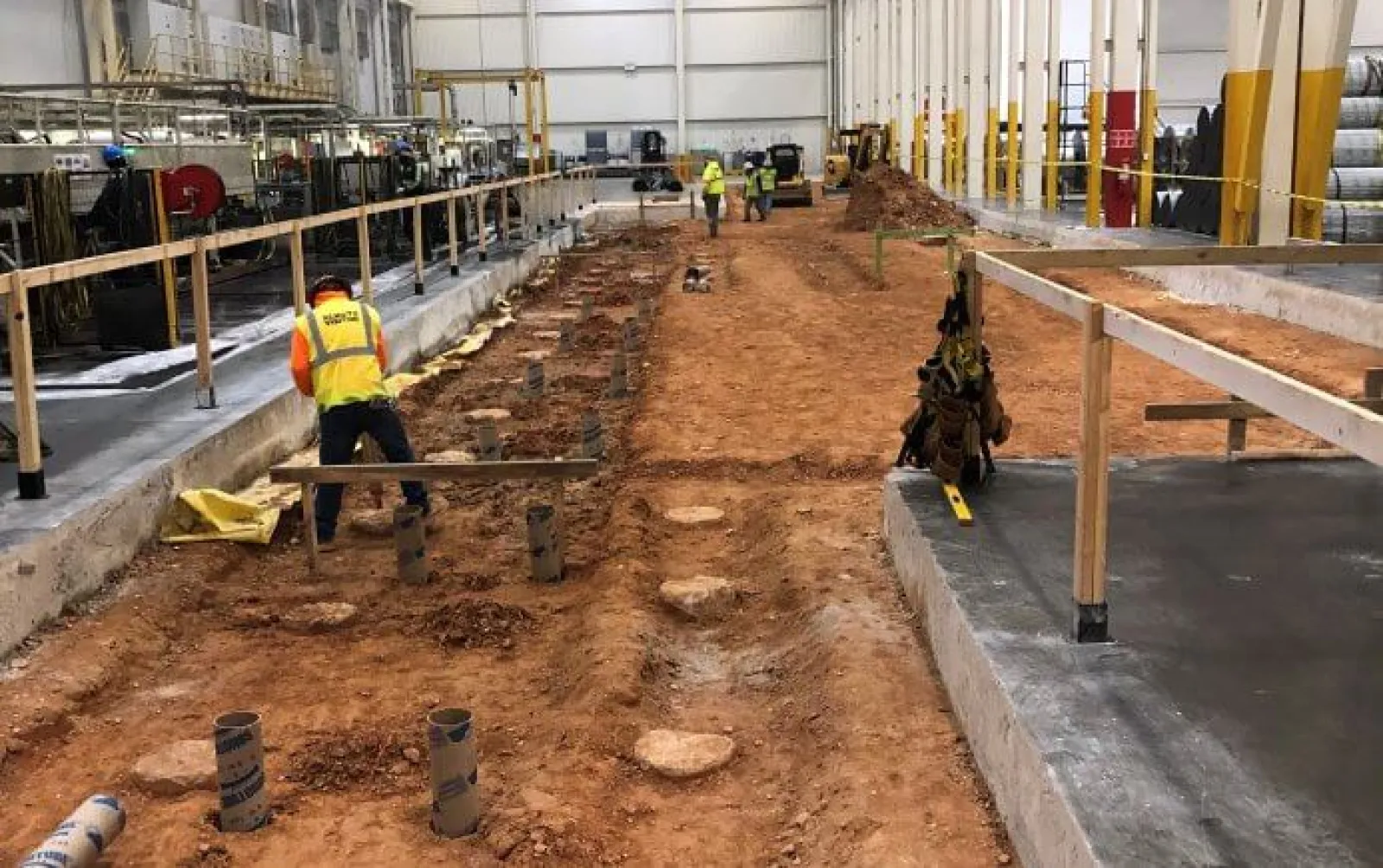 Construction workers preparing a large industrial site foundation with soil excavation and concrete flooring inside a warehouse.