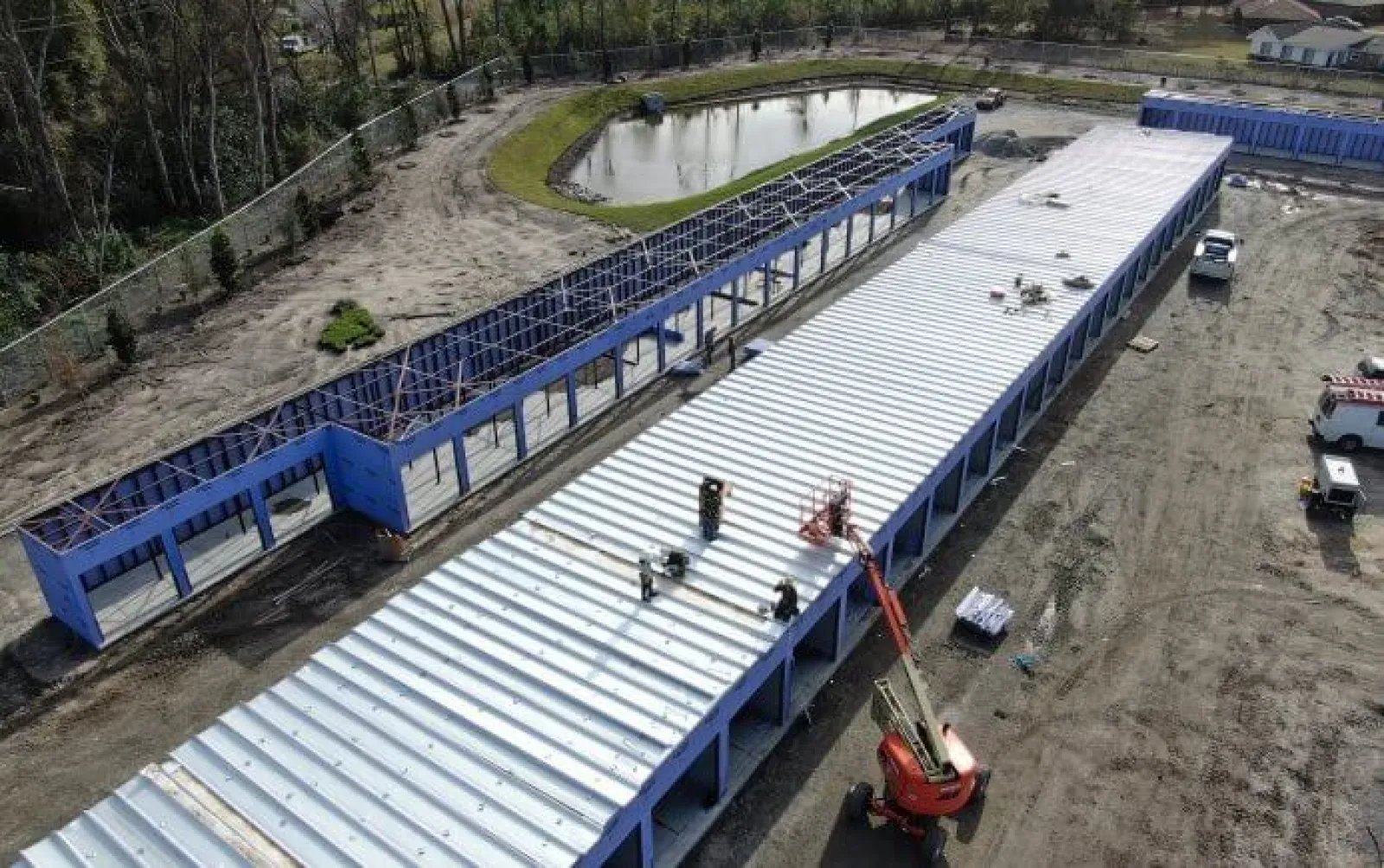 Aerial view of blue-framed industrial buildings under construction near a pond with workers on the metal roof.